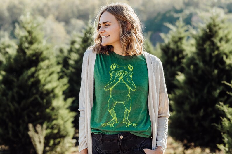 May include: A woman wearing a green t-shirt with a yellow outline of a frog covering its eyes with its hands. The woman is smiling and wearing a white cardigan.