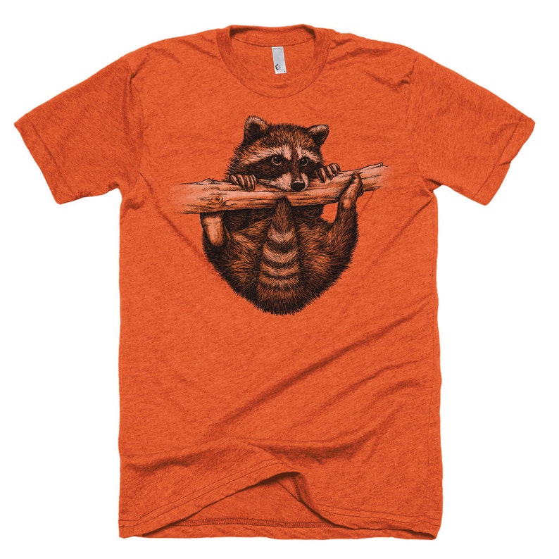 Puede incluir: Camiseta naranja con un gr&aacute;fico de un mapache colgado de una rama.