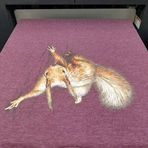 May include: A maroon t-shirt with a printed image of a squirrel peeking out from behind a tree branch.