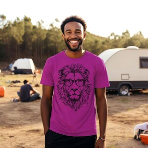 May include: A man wearing a pink t-shirt with a black and white illustration of a lion wearing glasses. The lion has a serious expression. The man is standing in front of a camper van and a tent.