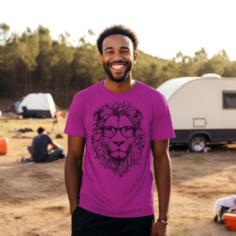 May include: A man wearing a pink t-shirt with a black and white illustration of a lion wearing glasses. The lion has a serious expression. The man is standing in front of a camper van and a tent.