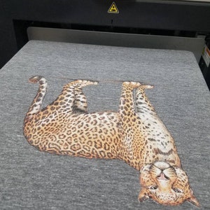 Puede incluir: Una camiseta gris con un estampado detallado de leopardo. El leopardo est&aacute; representado tumbado boca arriba, con la cabeza vuelta hacia el espectador. El estampado muestra el distintivo patr&oacute;n de manchas del animal.