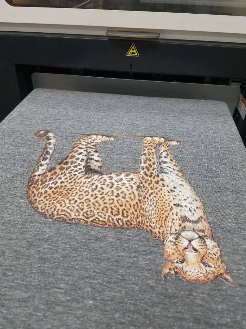 Puede incluir: Una camiseta gris con un estampado detallado de leopardo. El leopardo est&aacute; representado tumbado boca arriba, con la cabeza vuelta hacia el espectador. El estampado muestra el distintivo patr&oacute;n de manchas del animal.