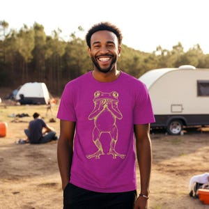 May include: A man wearing a magenta t-shirt with a yellow outline of a frog covering its face. The frog is standing on its hind legs with its arms covering its face.