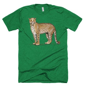 Puede incluir: Camiseta verde con una ilustraci&oacute;n detallada de un leopardo. El leopardo se representa en un estilo realista con un pelaje manchado de color marr&oacute;n y negro, de pie sobre una fina l&iacute;nea verde. La camiseta tiene cuello redondo y mangas cortas.