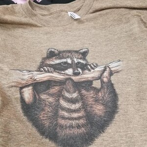 Puede incluir: Una camiseta marr&oacute;n con un estampado gr&aacute;fico de un mapache colgado de una rama.