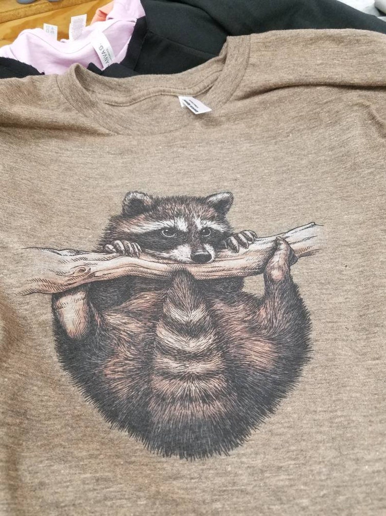 Puede incluir: Una camiseta marr&oacute;n con un estampado gr&aacute;fico de un mapache colgado de una rama.