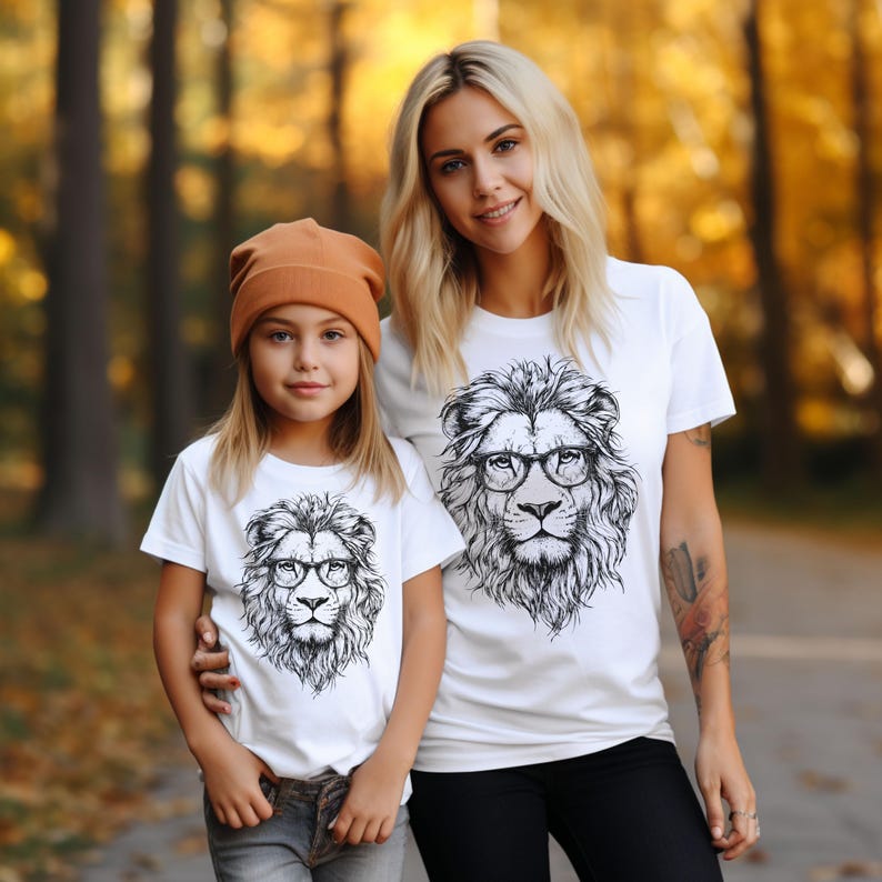 May include: Two people wearing white t-shirts with a black and white lion graphic wearing glasses. The lion graphic is centred on the shirts. The person on the left is a child wearing a brown beanie. The person on the right is an adult with blonde hair.