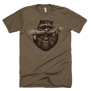Puede incluir: Una camiseta marr&oacute;n con un gr&aacute;fico de un mapache colgado de una rama. El mapache est&aacute; mirando hacia abajo con una expresi&oacute;n curiosa.