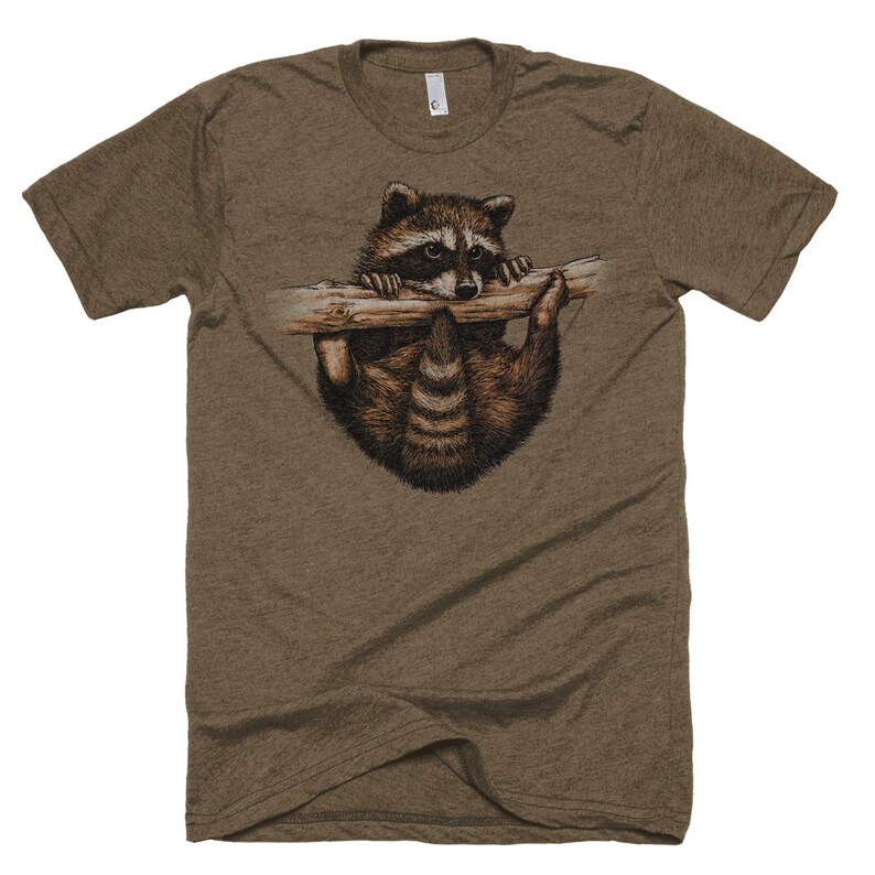 Puede incluir: Una camiseta marr&oacute;n con un gr&aacute;fico de un mapache colgado de una rama. El mapache est&aacute; mirando hacia abajo con una expresi&oacute;n curiosa.