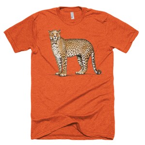 Puede incluir: Camiseta naranja de manga corta con una ilustraci&oacute;n detallada de un leopardo. El leopardo est&aacute; representado en un estilo realista con un pelaje manchado, de pie sobre una superficie blanca. La camiseta tiene cuello redondo y una textura jaspeada.