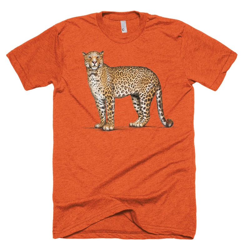 Puede incluir: Camiseta naranja de manga corta con una ilustraci&oacute;n detallada de un leopardo. El leopardo est&aacute; representado en un estilo realista con un pelaje manchado, de pie sobre una superficie blanca. La camiseta tiene cuello redondo y una textura jaspeada.