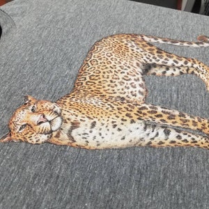 Puede incluir: Una camiseta gris jaspeado con una ilustraci&oacute;n detallada de un leopardo. El leopardo se representa en una pose relajada, con su pelaje manchado y sus rasgos faciales claramente visibles. La tela de la camiseta tiene un aspecto texturizado.