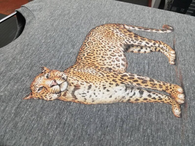 Puede incluir: Una camiseta gris jaspeado con una ilustraci&oacute;n detallada de un leopardo. El leopardo se representa en una pose relajada, con su pelaje manchado y sus rasgos faciales claramente visibles. La tela de la camiseta tiene un aspecto texturizado.
