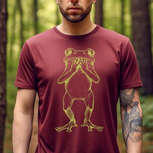 May include: A maroon t-shirt with a yellow outline of a frog covering its face with its hands.