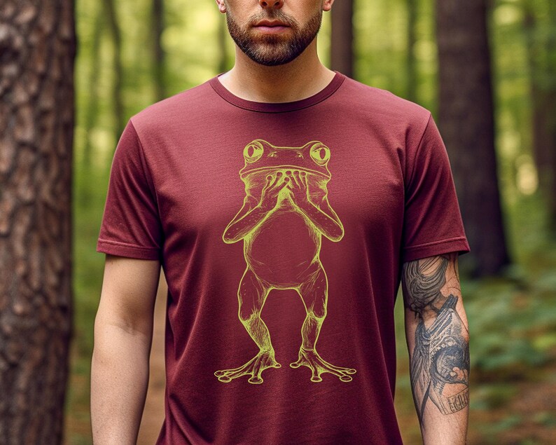 May include: A maroon t-shirt with a yellow outline of a frog covering its face with its hands.