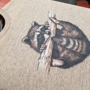 Puede incluir: Una camiseta gris con una ilustraci&oacute;n en blanco y negro de un mapache agarrado a una rama.