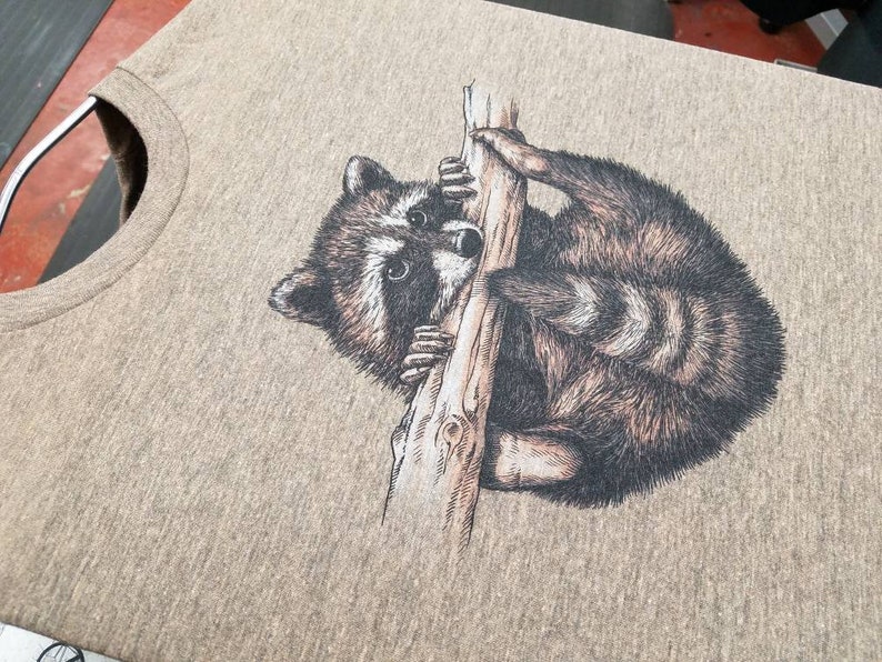Puede incluir: Una camiseta gris con una ilustraci&oacute;n en blanco y negro de un mapache agarrado a una rama.