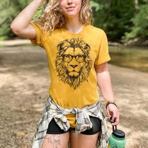 May include: A woman wearing a mustard yellow t-shirt with a black and white illustration of a lion wearing glasses. The lion is facing forward and has a mane of hair. The woman is wearing a plaid shirt tied around her waist and black shorts.