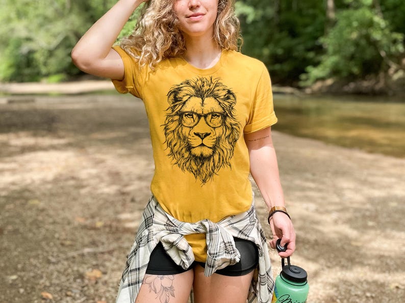 May include: A woman wearing a mustard yellow t-shirt with a black and white illustration of a lion wearing glasses. The lion is facing forward and has a mane of hair. The woman is wearing a plaid shirt tied around her waist and black shorts.