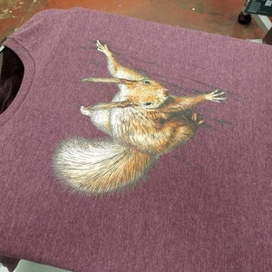 May include: A maroon t-shirt with a printed image of a squirrel climbing a tree.