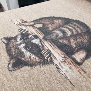 Puede incluir: Un mapache marr&oacute;n y negro colgado de una rama con sus patas agarrando la rama. El mapache est&aacute; mirando hacia la izquierda de la imagen.