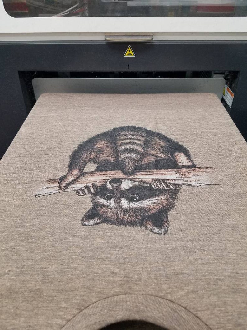 Puede incluir: Una camiseta gris con una imagen impresa de un mapache colgado boca abajo de una rama.