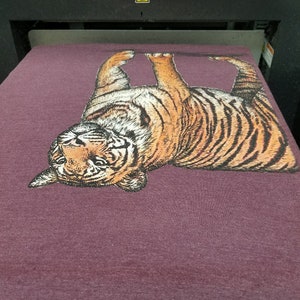 May include: A purple t-shirt with a printed image of a tiger. The tiger is facing the camera with its head tilted back and its mouth slightly open. The tiger is printed in shades of brown, orange, and black.