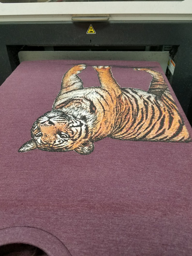 May include: A purple t-shirt with a printed image of a tiger. The tiger is facing the camera with its head tilted back and its mouth slightly open. The tiger is printed in shades of brown, orange, and black.