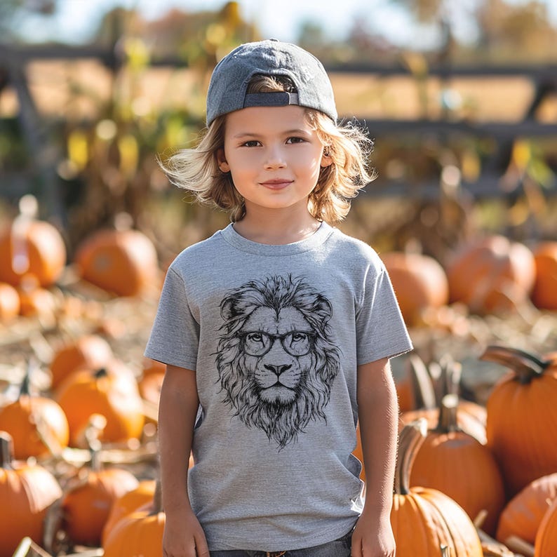 May include: A young child wearing a grey t-shirt with a black and white illustration of a lion wearing glasses. The child is also wearing a dark blue baseball cap. The child is standing in front of a pile of pumpkins.