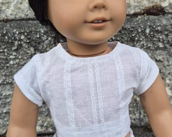 Peut inclure: Une poupée aux cheveux brun foncé et aux yeux bleus portant un haut blanc à manches courtes avec des détails en dentelle verticale et un pantalon blanc avec un motif ondulé texturé horizontal. La poupée a le teint clair.