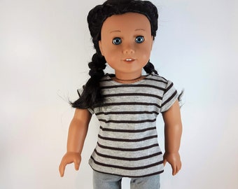 Peut inclure: Une poupée portant un t-shirt à manches courtes rayé gris et marron. La poupée a de longs cheveux noirs et des yeux bleus.