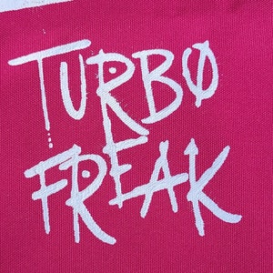 May include: A close-up of a magenta fabric with the words "TURBO FREAK" in white, handwritten-style lettering. The text is centered and takes up most of the frame, with a slightly distressed aesthetic.