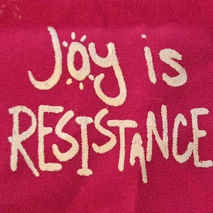 Peut inclure: Gros plan sur un tissu magenta avec l'inscription blanche "Joy is Resistance" dans une écriture manuscrite. Le texte est centré et occupe la majeure partie du cadre, les mots étant empilés verticalement.