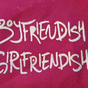 Puede incluir: Primer plano de una tela magenta con las palabras "BOYFRIENDISH" y "GIRLFRIENDISH" en blanco, con letras manuscritas. El texto está dispuesto en dos líneas, una encima de la otra.