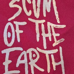 May include: A close-up of a magenta fabric with the words "SCUM OF THE EARTH" in a distressed, white, hand-drawn font. The text is centered and takes up most of the frame, with the letters slightly faded.