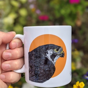 May include: White ceramic mug with a black and white illustration of a falcon on an orange circle. The mug has a black handle.