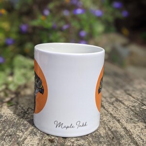 May include: White ceramic mug with an orange circle and a black and white bird illustration. The mug has the text "Maple Inkk" printed on the bottom.