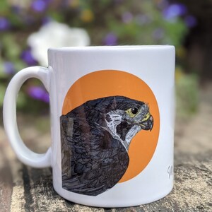 May include: White ceramic mug with a black and white illustration of a bird of prey on an orange circle. The bird is facing right and has a yellow eye.