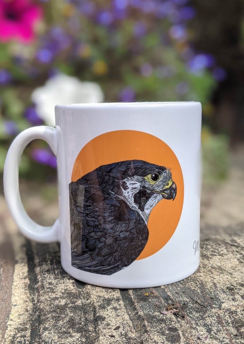 May include: White ceramic mug with a black and white illustration of a bird of prey on an orange circle. The bird is facing right and has a yellow eye.
