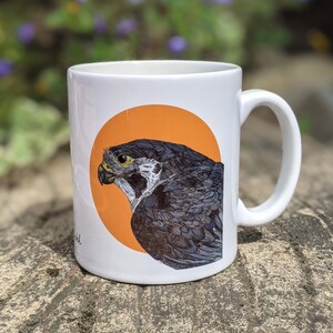 May include: White ceramic mug with a black and gray falcon illustration on an orange circle. The falcon is facing left and has a yellow eye.