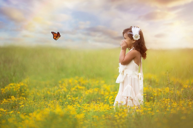 Puede incluir: Una ni&ntilde;a peque&ntilde;a con un vestido blanco y una diadema de plumas sonr&iacute;e mientras observa a una mariposa volar en un campo de flores amarillas.
