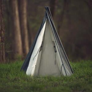 May include: A white and grey teepee tent set up in a grassy area with a blurred background of trees.