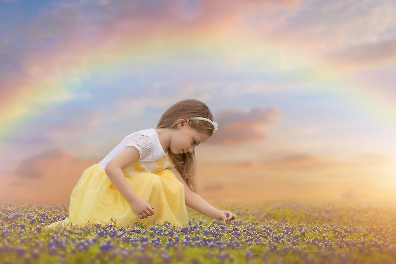 Puede incluir: Una ni&ntilde;a peque&ntilde;a con un vestido amarillo con ribetes de encaje blanco est&aacute; arrodillada en un campo de flores azules. Est&aacute; llegando para tocar una flor con su mano derecha. El cielo detr&aacute;s de ella es de un suave rosa pastel y azul con un arco&iacute;ris tenue.