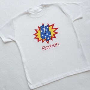 May include: White t-shirt with a blue and white polka dot number four in a red and yellow comic book style explosion. The name Roman is embroidered below the number.