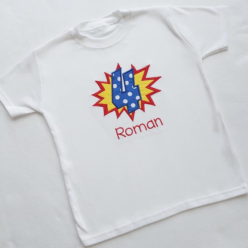 May include: White t-shirt with a blue and white polka dot number four in a red and yellow comic book style explosion. The name Roman is embroidered below the number.