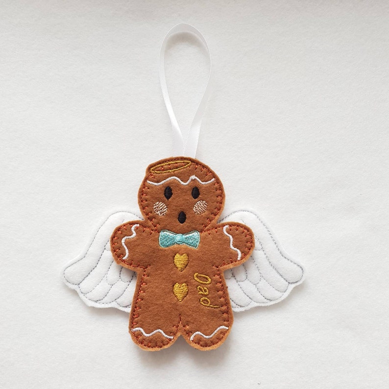 Puede incluir: Adorno de hombre de jengibre marr&oacute;n con alas de &aacute;ngel blancas y una pajarita azul. El adorno tiene un halo dorado y la palabra "Dad" bordada en el cuerpo. Se adjunta una cinta blanca para colgarlo.