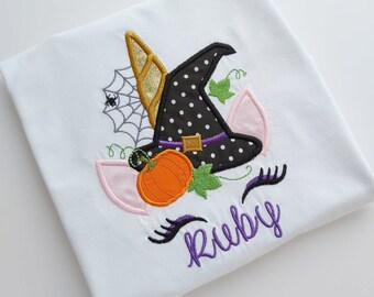 Puede incluir: Una camiseta blanca con un diseño bordado de un sombrero de bruja negro de lunares con una banda morada, un cuerno dorado, una calabaza naranja brillante, una telaraña con una araña, hojas verdes, orejas rosas y pestañas negras sobre el nombre Ruby.
