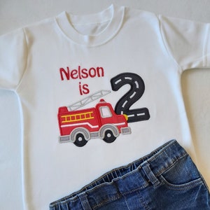 May include: White t-shirt with red embroidered text "Nelson is 2" and a red fire truck with a black number 2 behind it.
