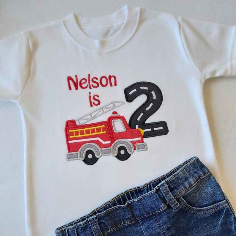 May include: White t-shirt with red embroidered text "Nelson is 2" and a red fire truck with a black number 2 behind it.